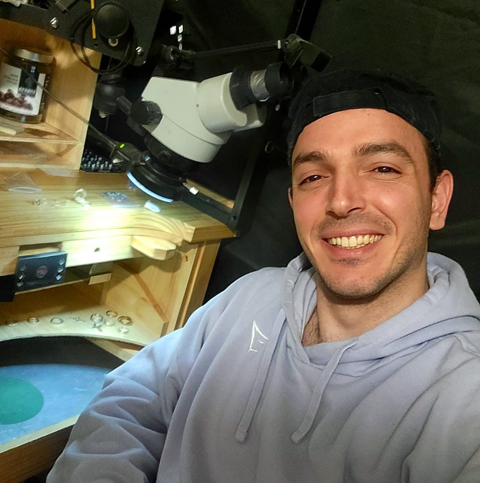 Dimitrios Koukoulis, the artisan behind Dimitrios Creations, smiling at his jewelry workstation with a microscope and tools, showcasing his dedication to craftsmanship and precision.