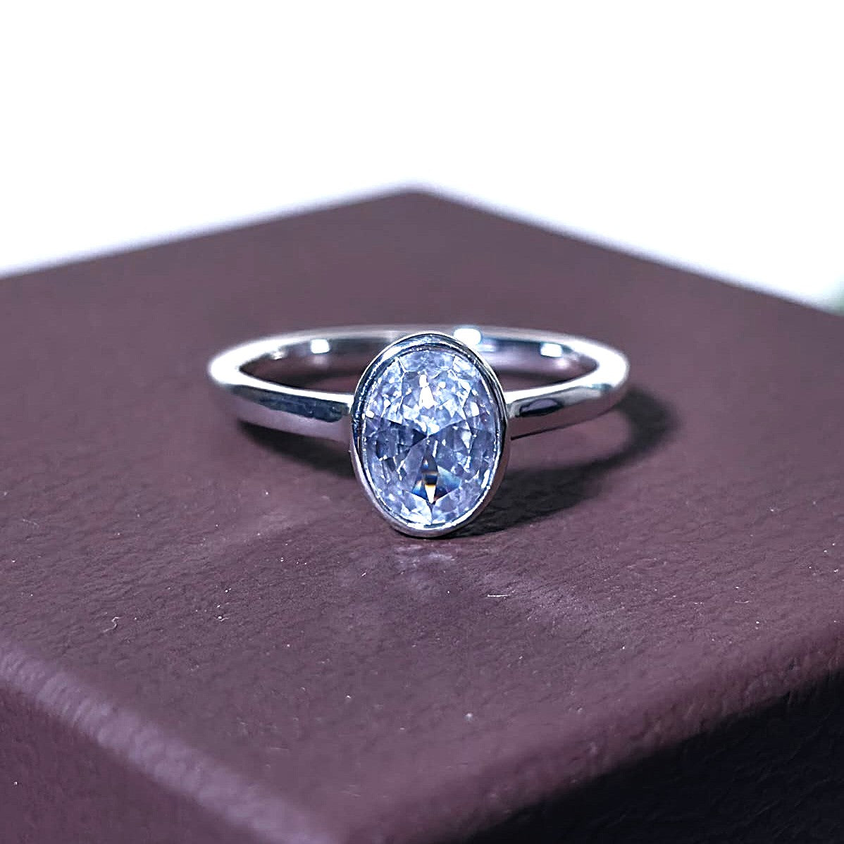 Close-up of a 1 ct oval lab-grown diamond engagement ring in 14K bezel setting on brown textured background.
