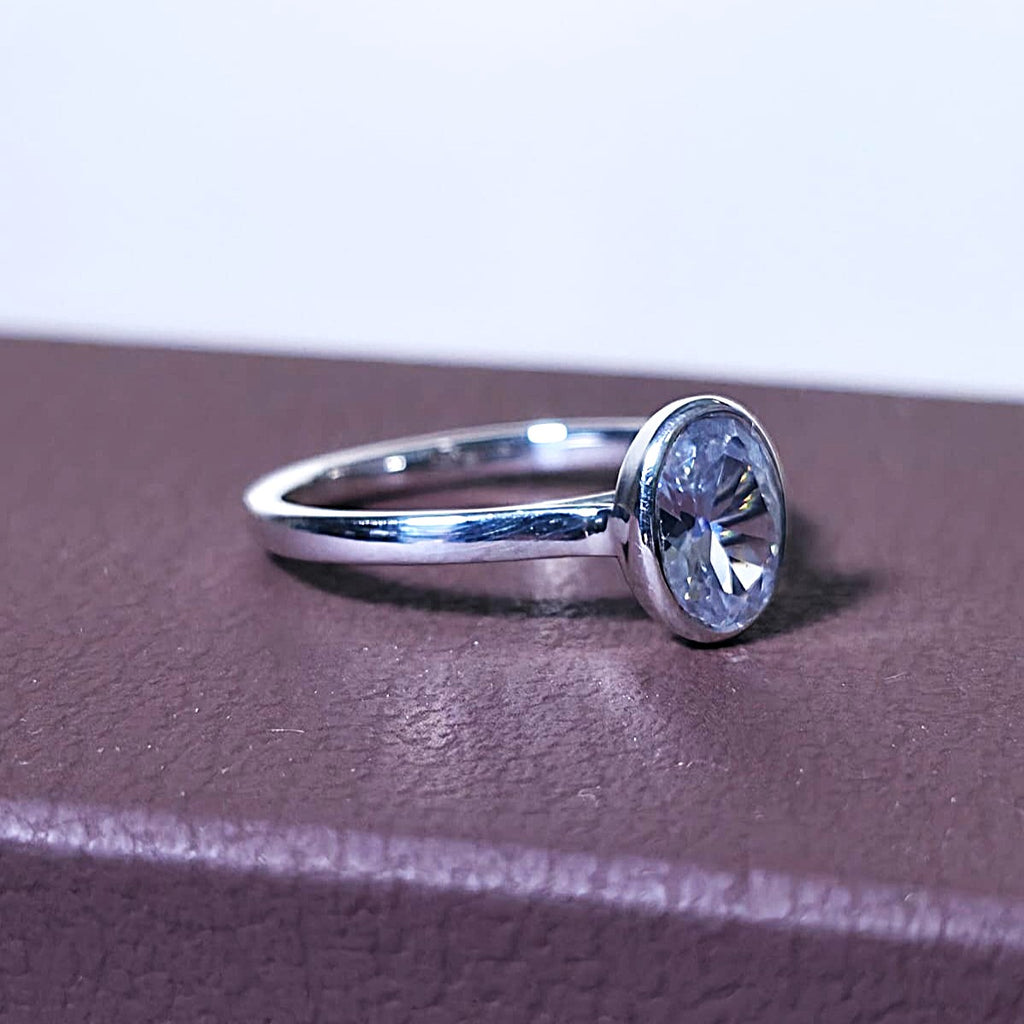 Close-up of 1 ct oval bezel engagement ring in 14K gold on a textured brown surface.