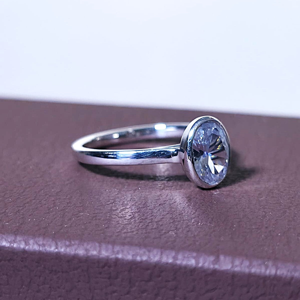 Close-up of 1 ct oval bezel engagement ring in 14K gold on a textured brown surface.