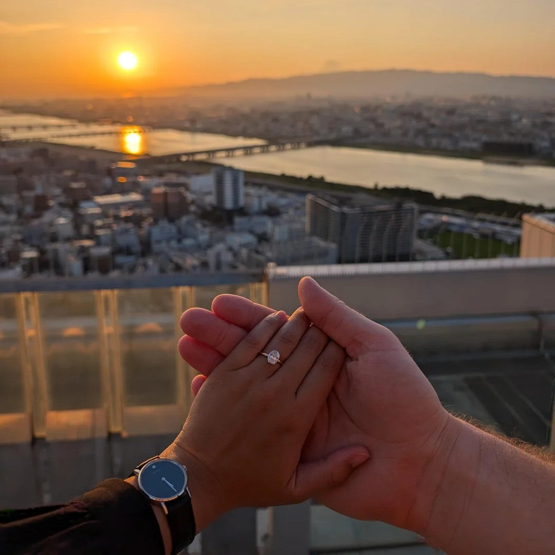 Close-up of hand with oval bezel engagement ring held against a sunset skyline backdrop.