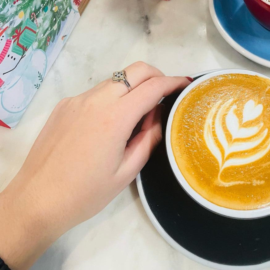Hand holding a cup of coffee with latte art on a marble surface wearing tyche clover ring